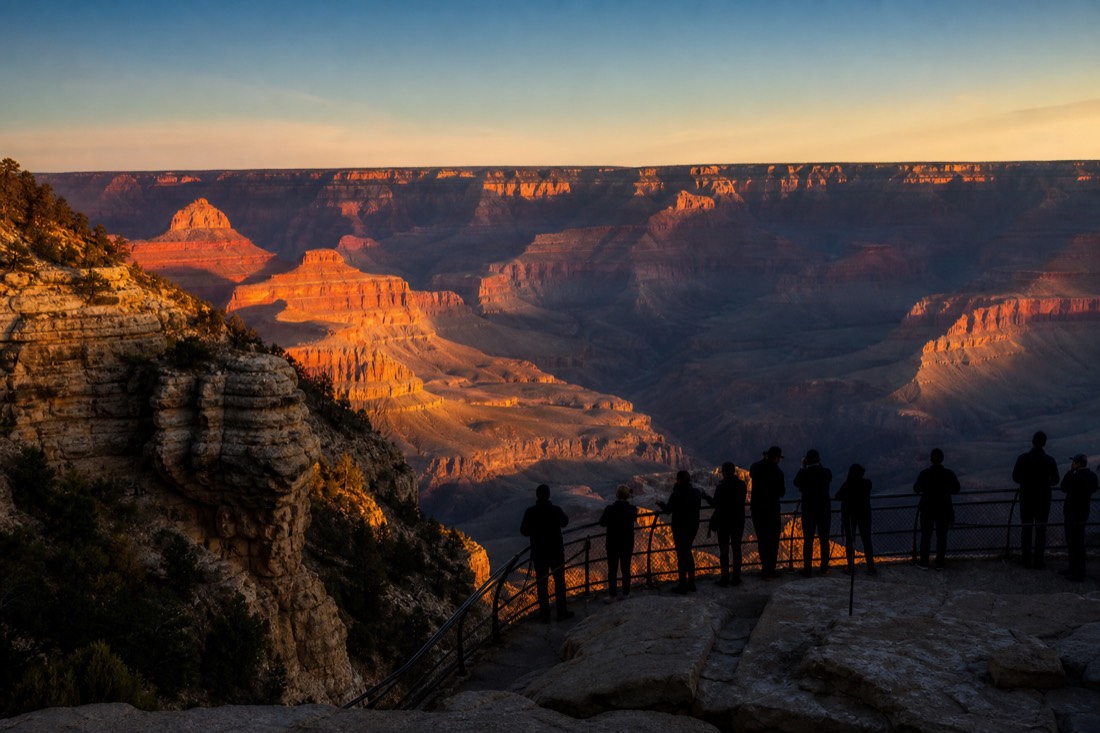 Silhouetted visitors at the Mather Point railing watching dawn color spill across the canyon buttes