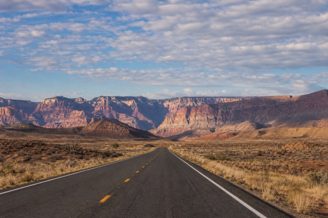 A desert highway running toward distant canyon country under a huge morning sky, the road empty in both directions