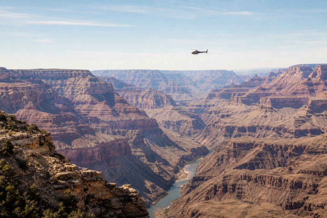 A helicopter crossing the Grand Canyon rim with the Colorado River winding through the gorge beneath
