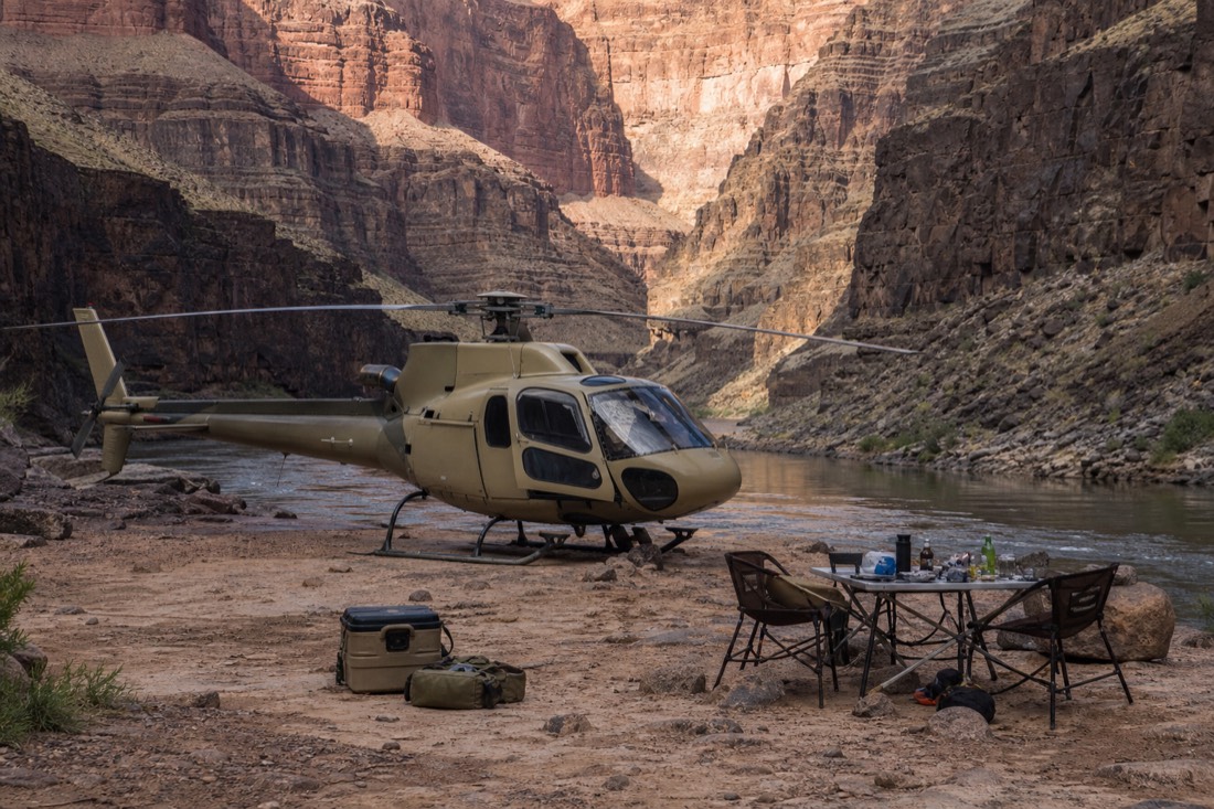 A helicopter parked on a canyon-floor landing pad while passengers look out toward the river