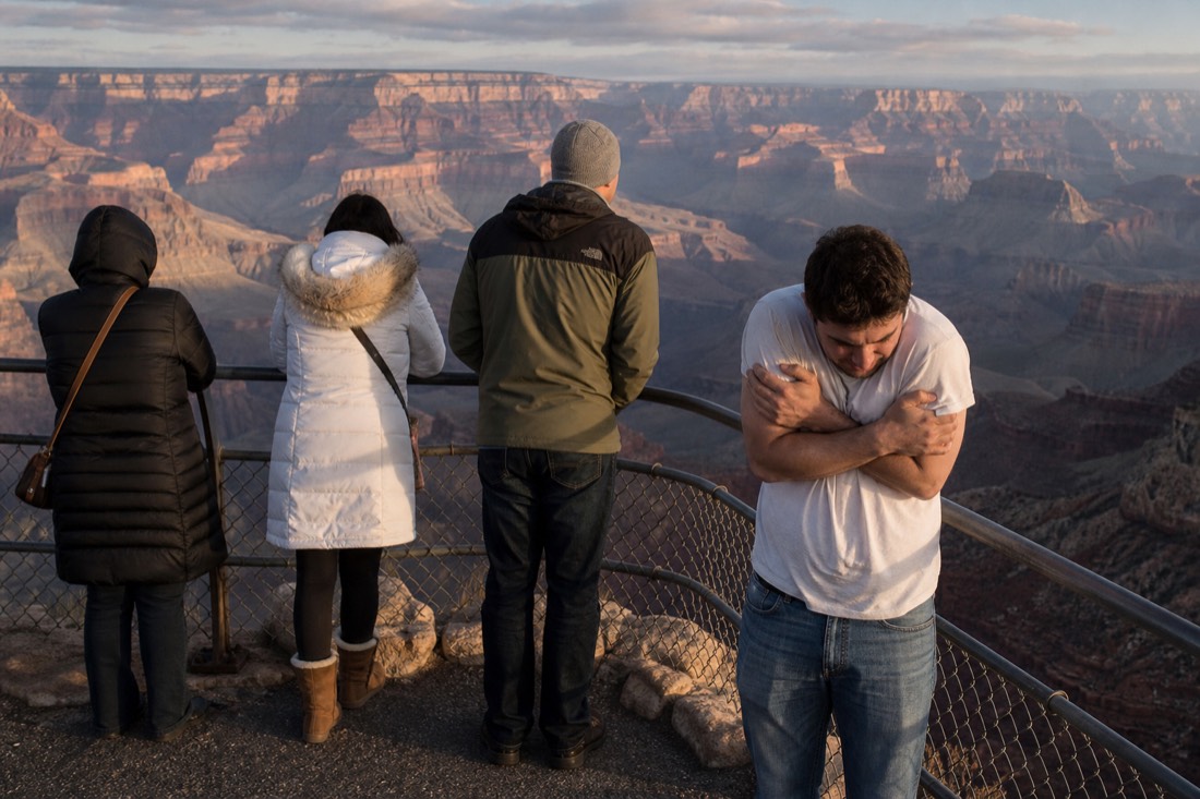 A visitor in a thin t-shirt hunched against cold wind at a Grand Canyon railing while others nearby wear winter coats