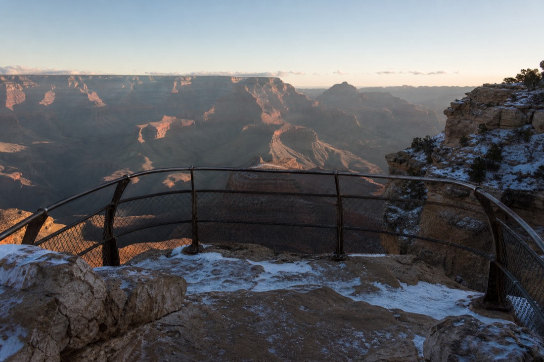 An empty snow-edged viewpoint railing above the Grand Canyon under low winter sun with no visitors in sight