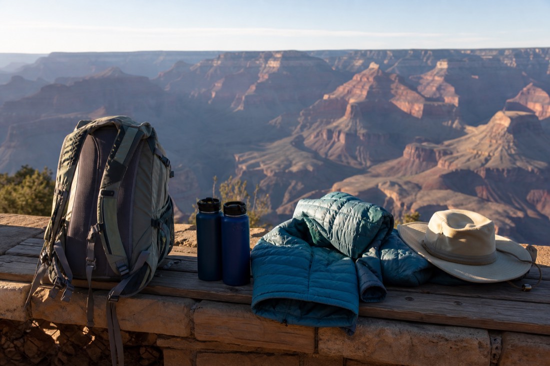 A daypack, warm jacket, water bottles and sun hat laid out on a wooden bench at a Grand Canyon overlook