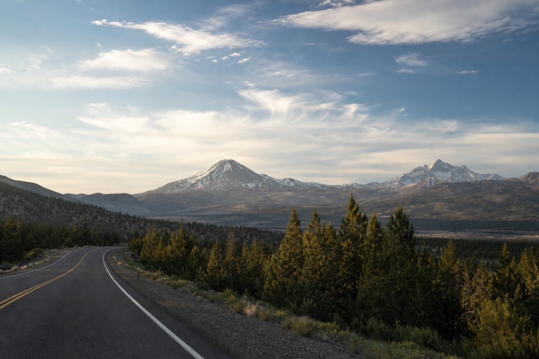 A forest highway curving northwest away from snow-dusted volcanic peaks under a wide morning sky