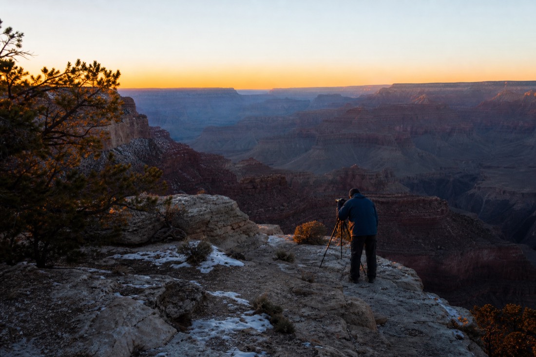 First light striking the canyon rim with a lone photographer setting up a tripod in the cold blue dawn