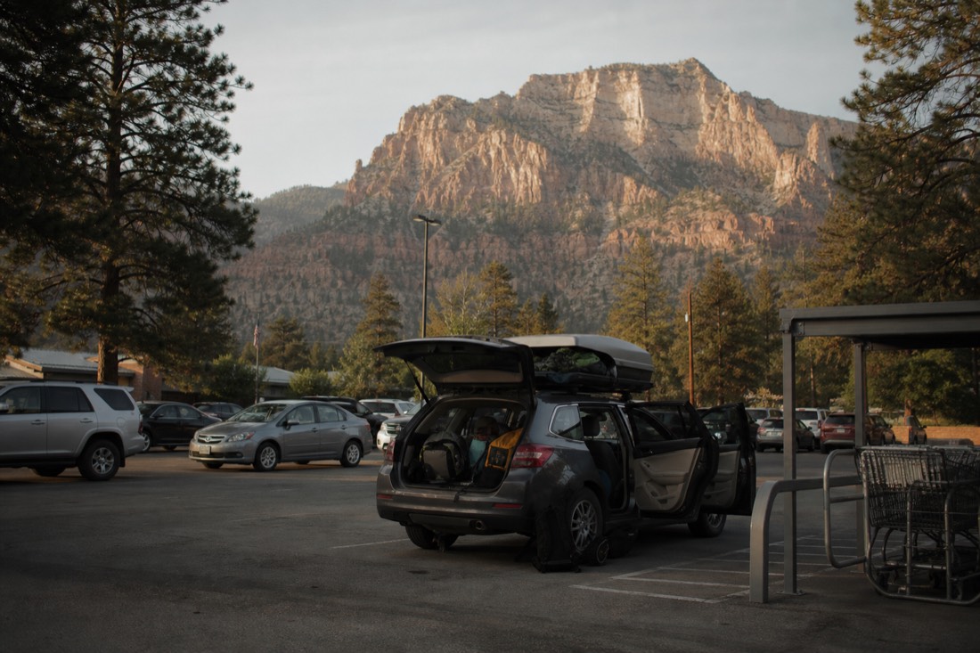 A supermarket car park in a pine-ringed mountain town at dawn with a loaded car and travellers stowing bags