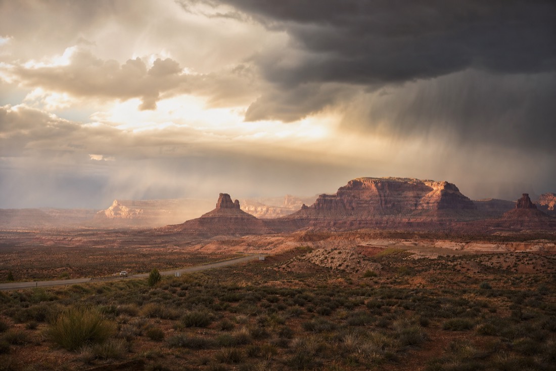A rain squall crossing distant desert mesas with sunlight breaking through the cloud edge above a highway