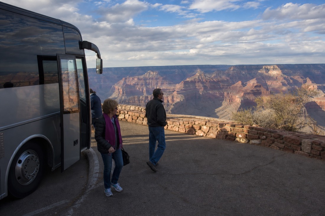 Passengers stepping down from a coach at a canyon viewpoint car park with the rim visible beyond the barrier