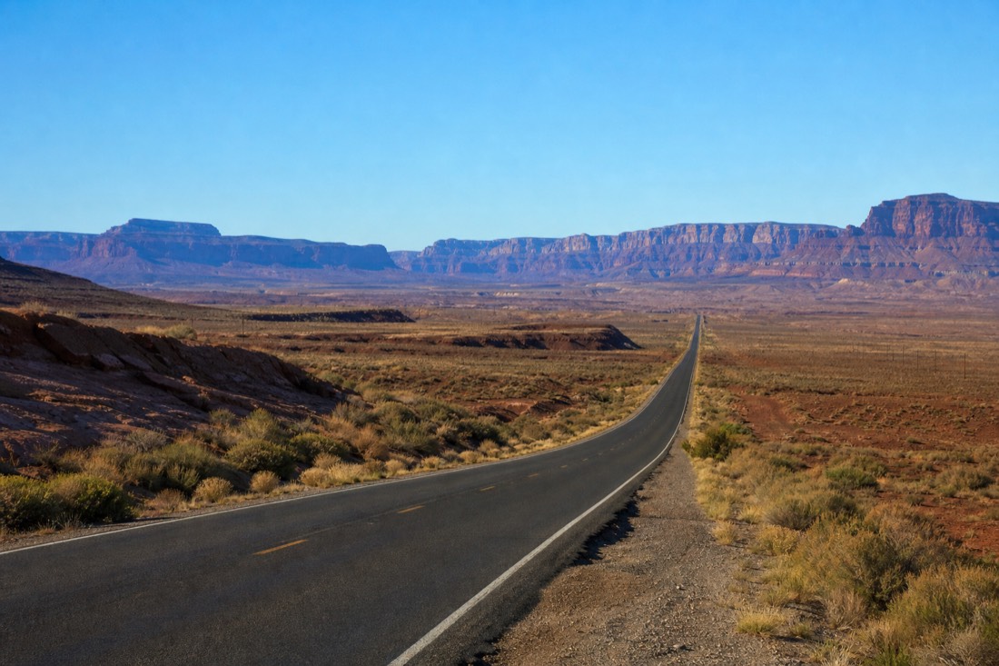 A long straight desert highway crossing empty scrubland with distant blue mountains under a hard midday sky