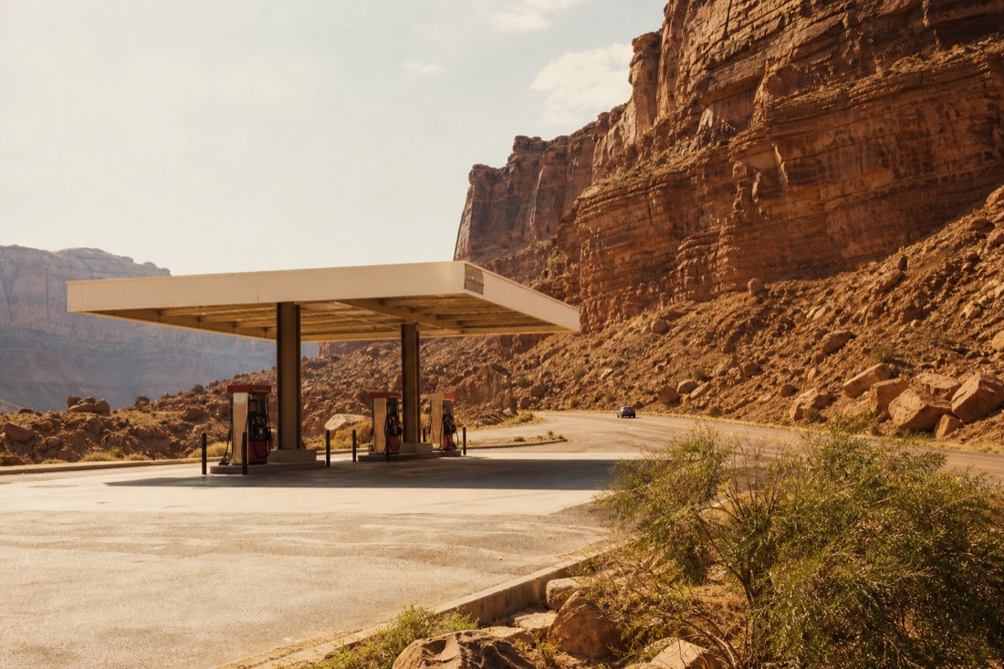 A lone fuel station with a shaded canopy beside a desert highway, heat haze rising off the forecourt