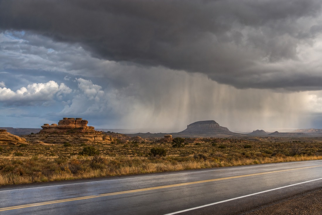 Dark monsoon clouds and rain curtains sweeping over high desert grassland with a wet highway in the foreground