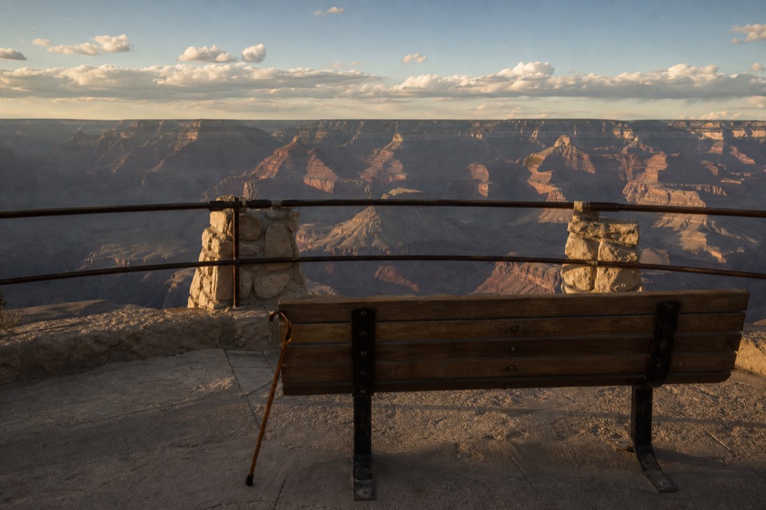 A traveler with a walking cane resting on a bench at a railed South Rim overlook in late afternoon light