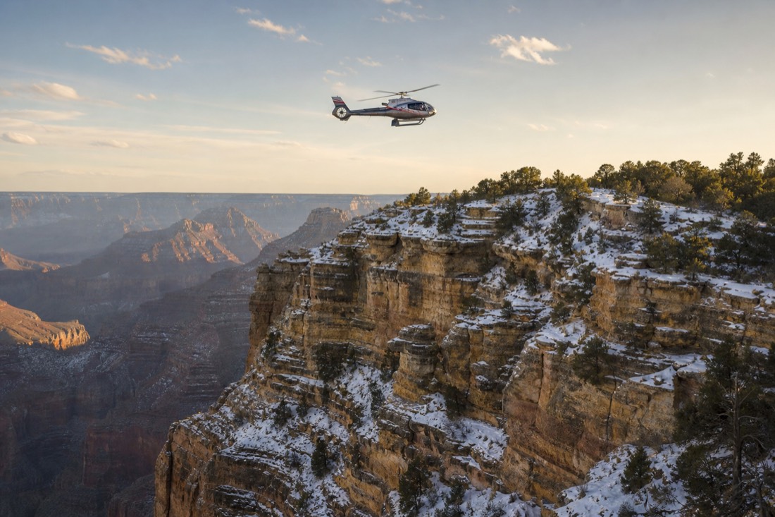 A tour helicopter crossing above a snow-dusted canyon rim in low winter sunlight