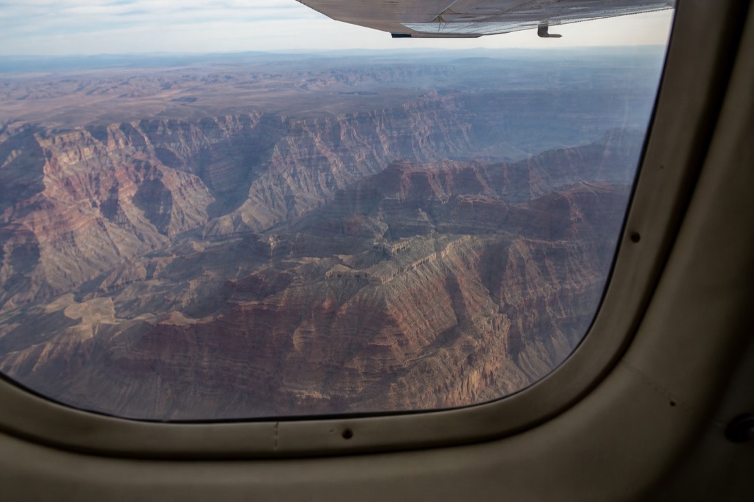 Interior view through a large curved aircraft window showing the layered canyon walls below