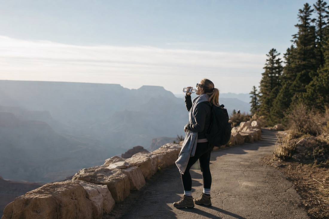 A hiker pausing to drink water on a paved rim path with pine forest and thin high-altitude light around them