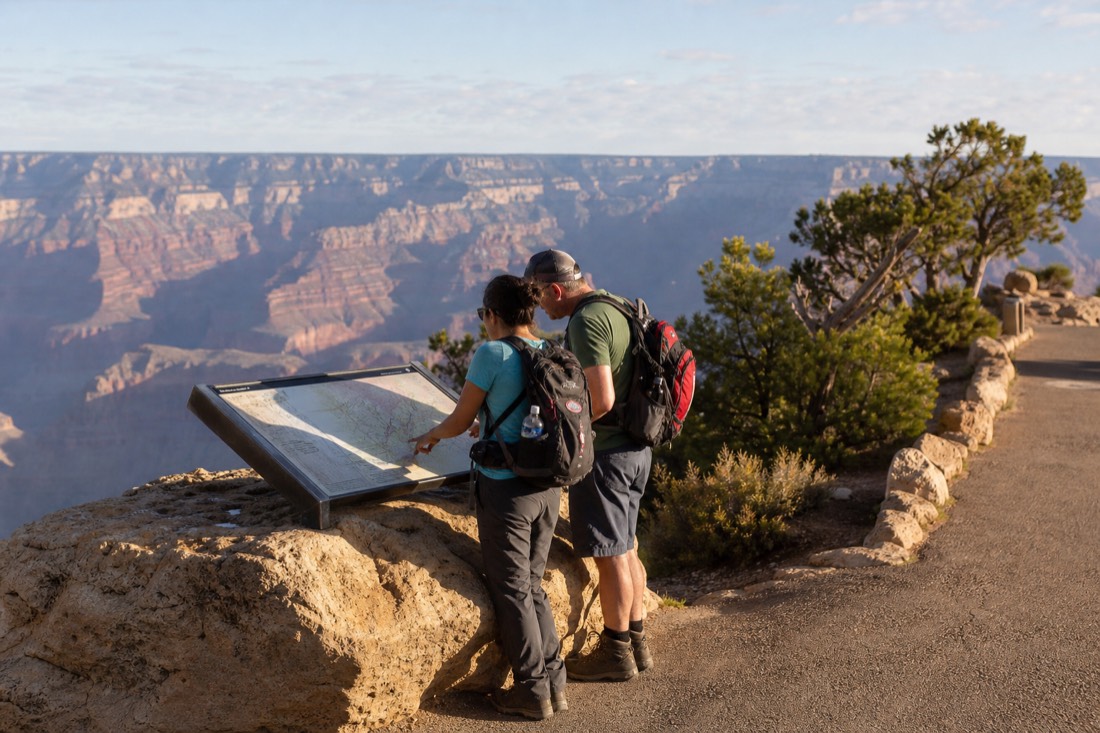 Travelers with daypacks studying a trail map board near the Grand Canyon visitor center in morning light
