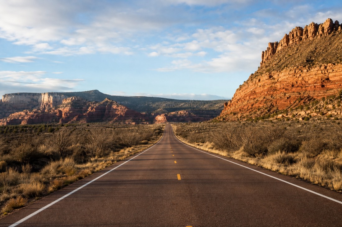 An empty two-lane Arizona highway running straight toward distant pine ridges under a wide morning sky