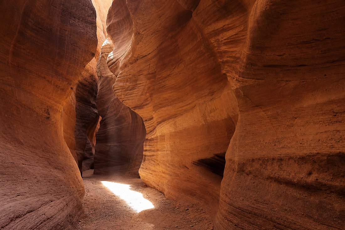 Sunlit sandstone walls curving overhead inside a narrow Arizona slot canyon with a shaft of light on the floor