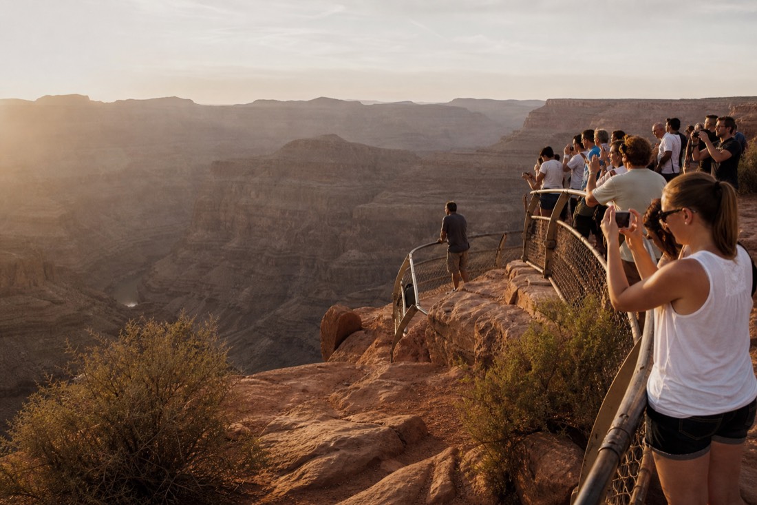 Visitors crowding a canyon railing taking photos while one person stands apart looking out at the gorge