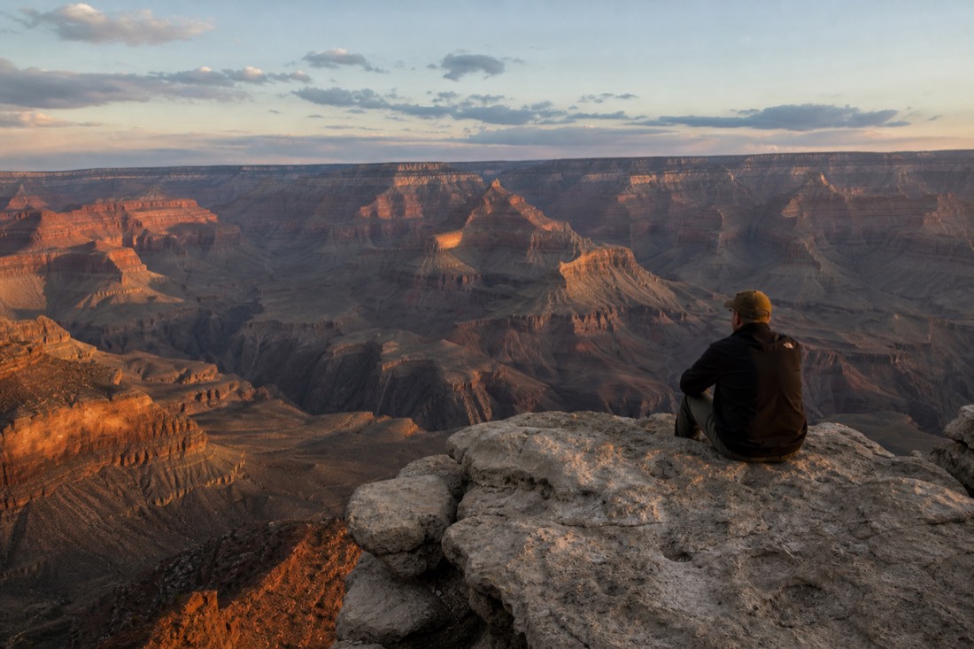 A visitor sitting still on a rock ledge above the Grand Canyon watching the light change with no one else nearby