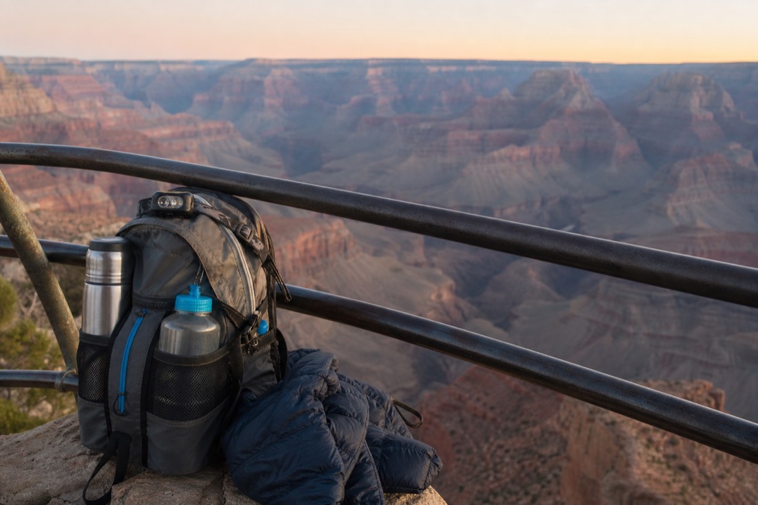 A small daypack with a headlamp, thermos, water bottles and a folded jacket beside a canyon overlook railing at dawn