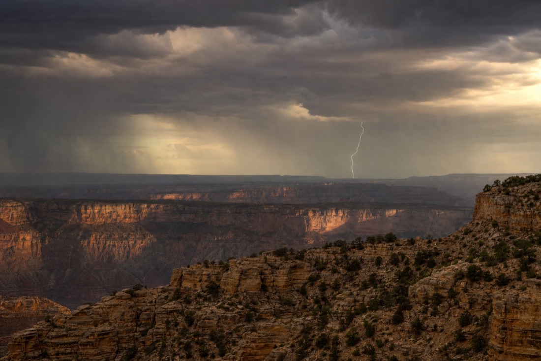 Lightning striking beyond a distant canyon rim under a bruised late-afternoon monsoon sky