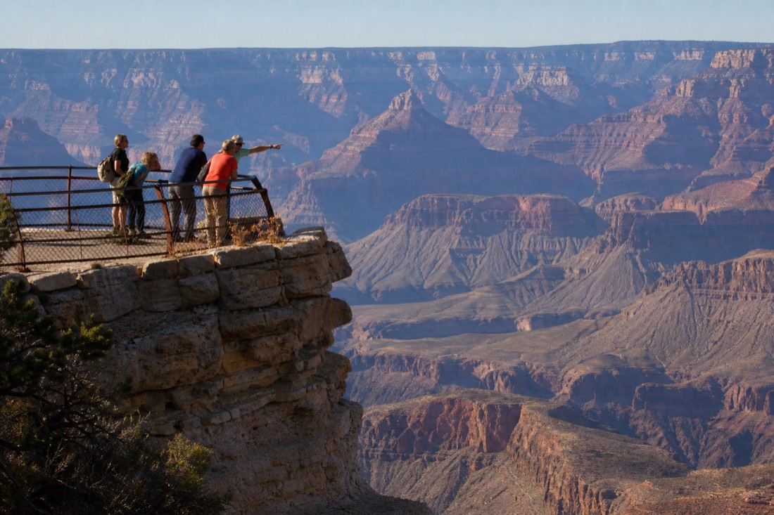 Small group of visitors leaning on a rim railing pointing across the canyon in mid-morning light