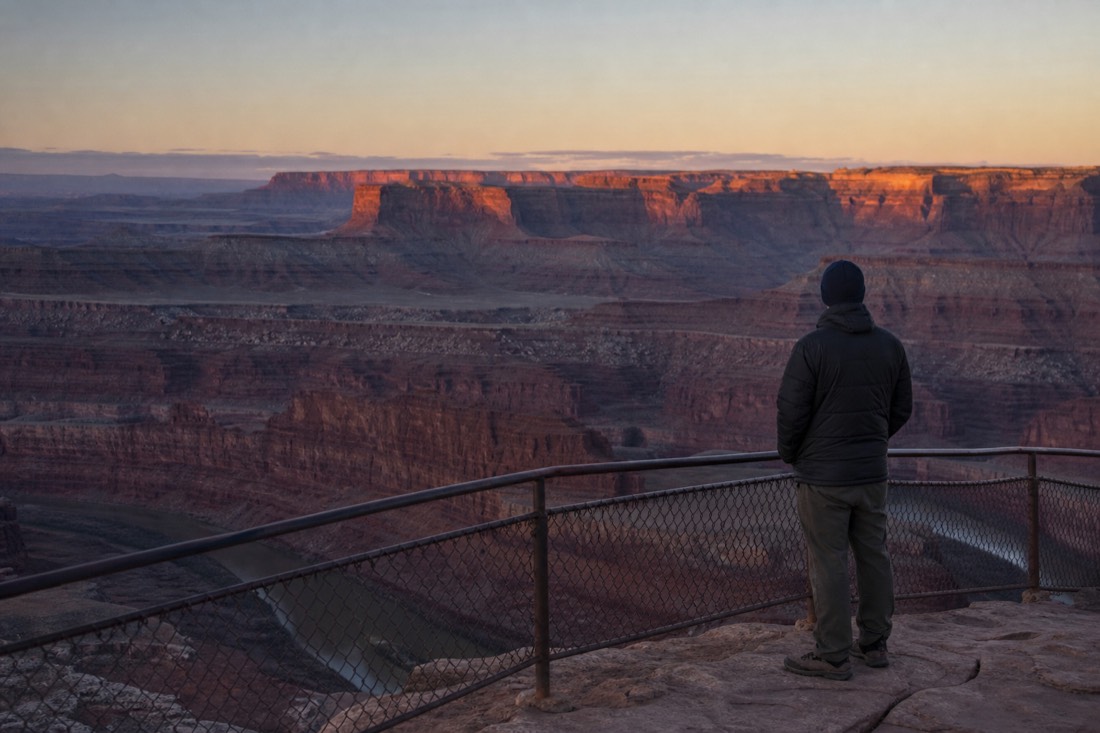Lone figure standing at a canyon railing before dawn as the first light reaches the far buttes