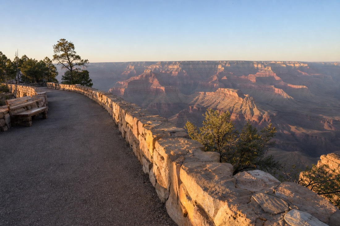 Wide paved rim path with a low stone wall and benches overlooking the canyon on a clear morning