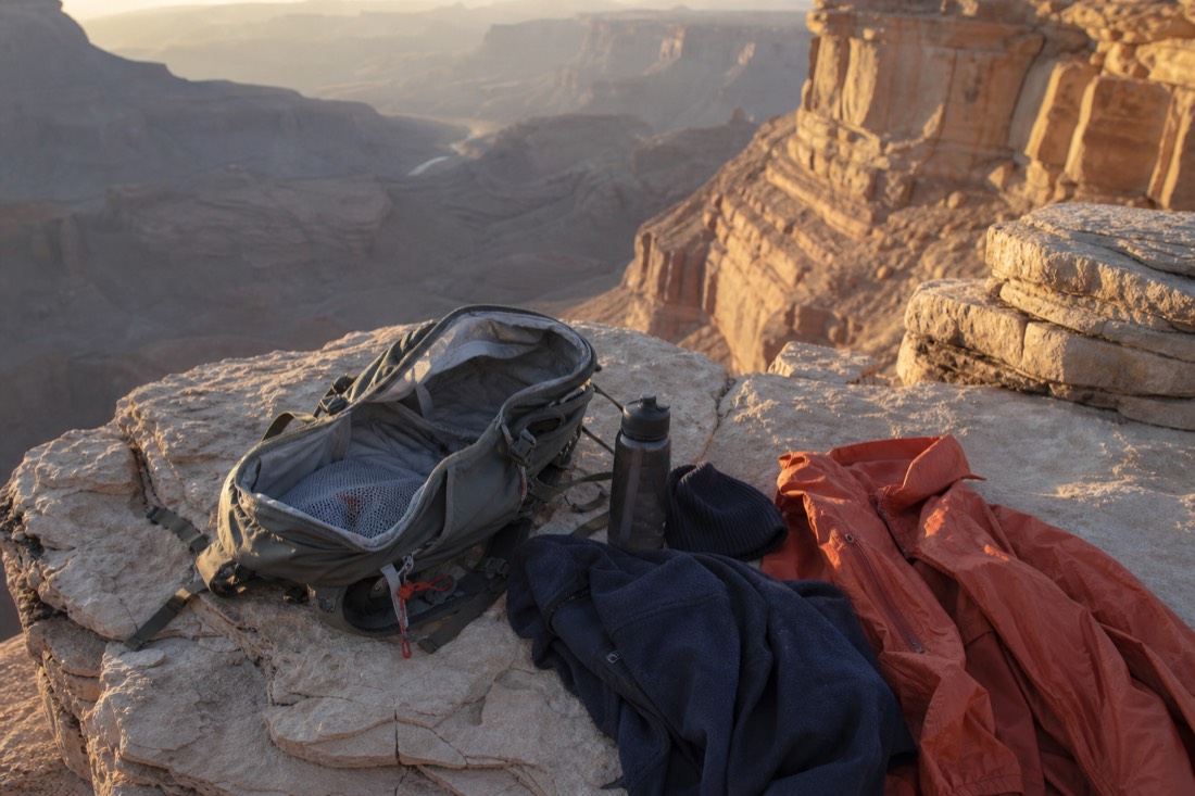 Open daypack on a rock ledge with a fleece, water bottle, hat and rain shell laid out beside it