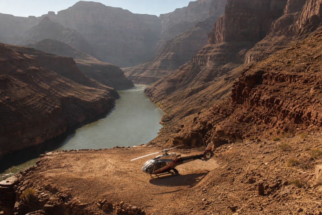 Small helicopter sitting on a dirt landing pad deep in the canyon beside the green Colorado River