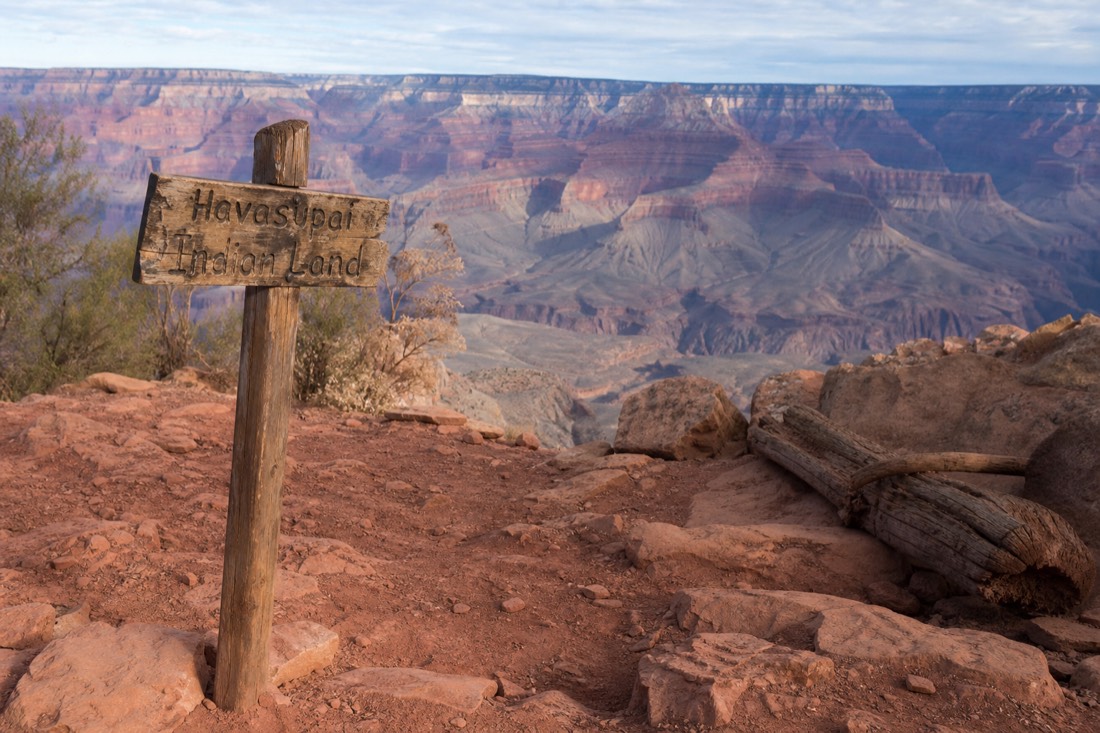 Weathered wooden signpost on red desert ground marking tribal land with the canyon rim beyond