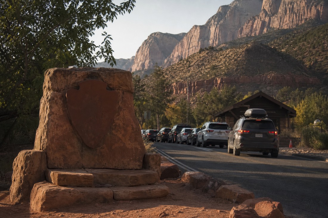 Park entrance sign and ranger kiosk in morning shade with a line of cars waiting on the two-lane road