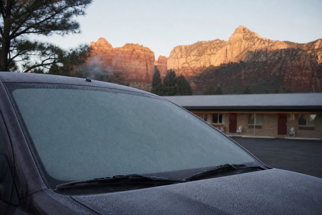 Frost on a car windscreen at dawn outside a pine-shaded motel with breath visible in the cold air