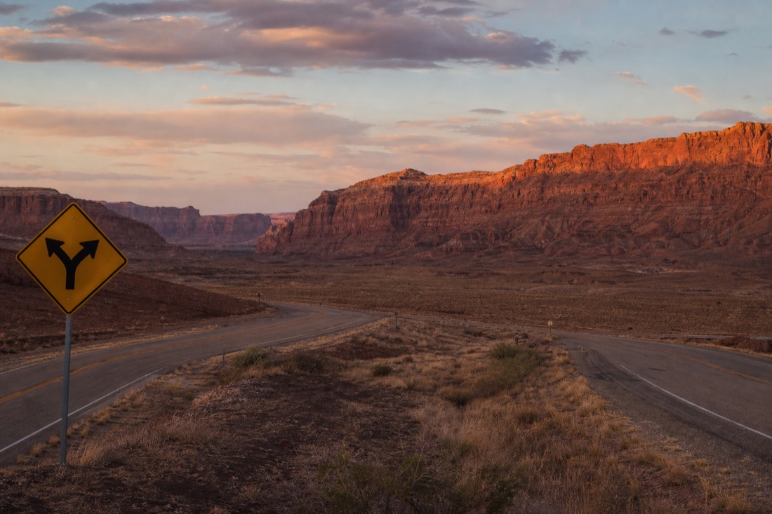 Highway junction sign at dusk in high desert with two roads splitting toward distant mesas
